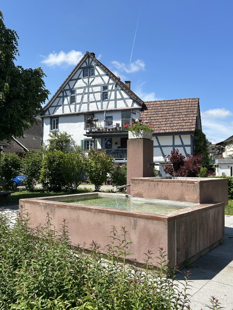 Moderner Brunnen vor altem Riegelbau